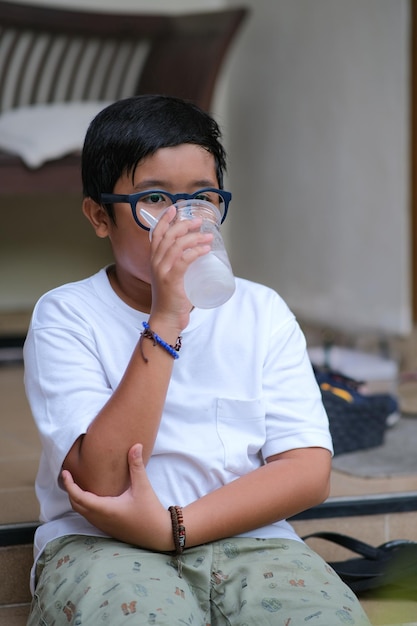 Um menino gordinho usando óculos sentado sozinho bebendo limonada gelada de um copo de plástico