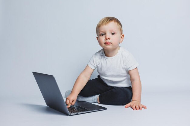 Um menino inteligente de 34 anos senta-se com um laptop em um fundo branco Uma criança em uma camiseta branca e calça preta se senta em um laptop e olha para a câmera Crianças modernas