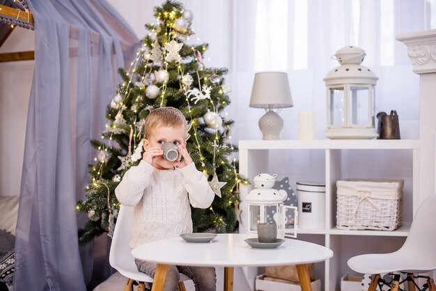 Um menino joga chá, bebe cacau em uma xícara cinza. Ao fundo há uma árvore de Natal