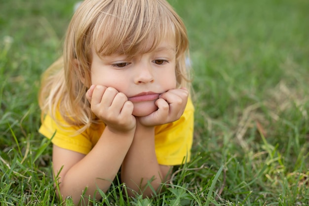 um menino loiro com cabelo comprido e uma misteriosa expressão pensativa no rosto está em um gramado verde