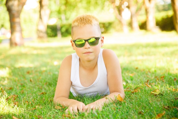 Um menino loiro com uma camiseta branca e óculos de sol está deitado na grama do parque