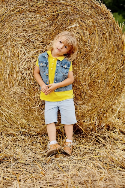 Um menino loiro com uma jaqueta amarela um colete jeans e shorts de linho está de pé ao lado de um palheiro