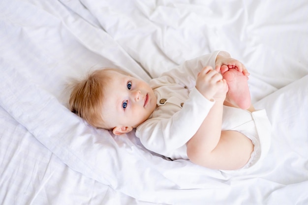 Um menino loiro de 6 meses brinca com as pernas em uma cama branca em um quarto brilhante depois de dormir de manhã em um bodysuit de algodão vista superior conceito de bens infantis