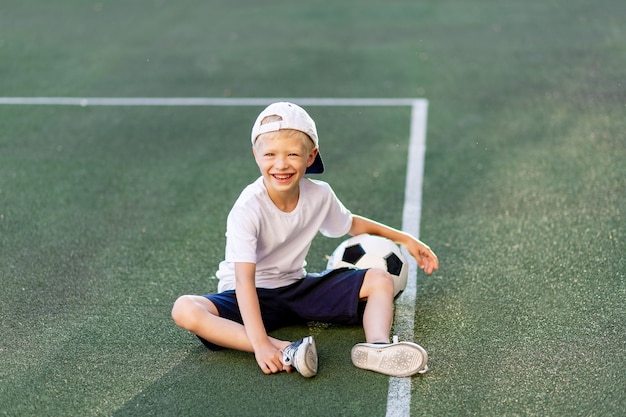 Um menino loiro de boné e uniforme esportivo sentado em um campo de futebol com uma bola de futebol