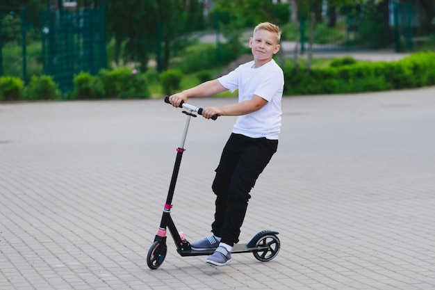 Um menino loiro de camiseta branca e calça preta andando de scooter. Foto de alta qualidade
