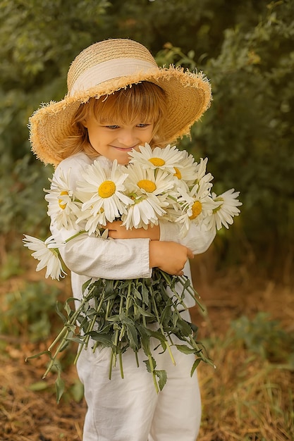 um menino loiro em um terno de linho branco e um chapéu de palha tem um buquê de margaridas nas mãos