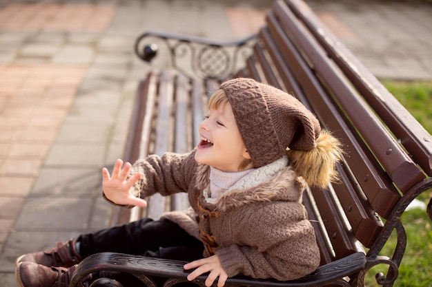 um menino loiro feliz em roupas marrons de malha está sentado em um banco do parque