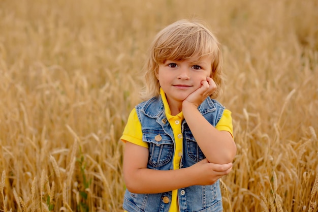 um menino loiro feliz em uma jaqueta amarela e um colete jeans está parado em um campo de trigo