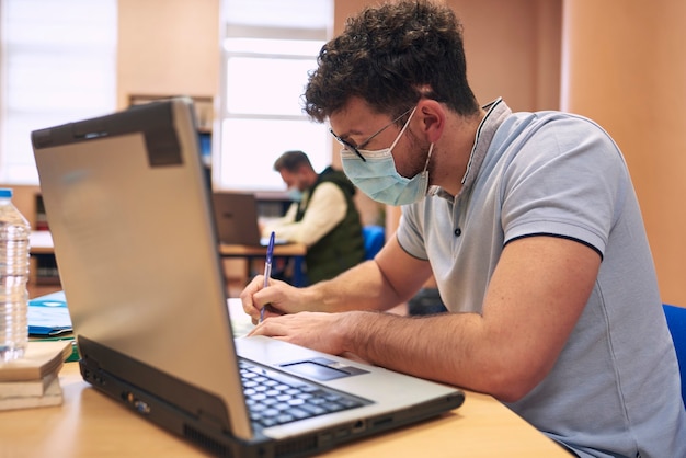 Foto um menino mascarado está estudando com um laptop na biblioteca