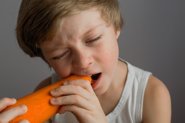 Um menino mordisca uma grande cenoura laranja em um cinza