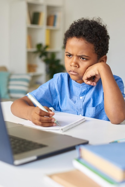 Foto um menino negro carrancudo e pensativo com cabelo encaracolado sentado à mesa e ouvindo o tutor durante uma videoconferência