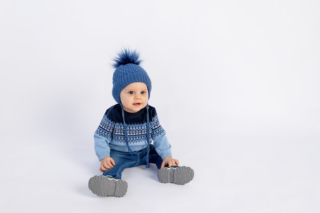 Um menino pequeno com um chapéu quente de inverno com um pompom e uma jaqueta azul sentado no branco