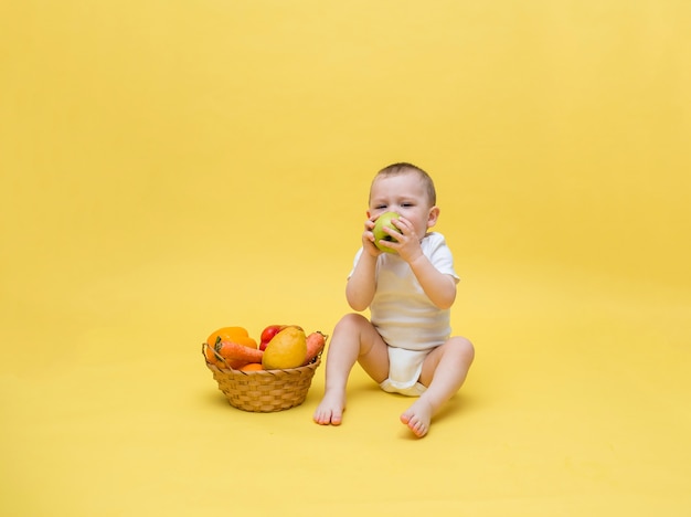 Um menino pequeno com uma cesta de vime com legumes e frutas em um espaço amarelo. O garoto está sentado em uma roupa branca e comendo uma maçã. Espaço livre em um espaço amarelo.