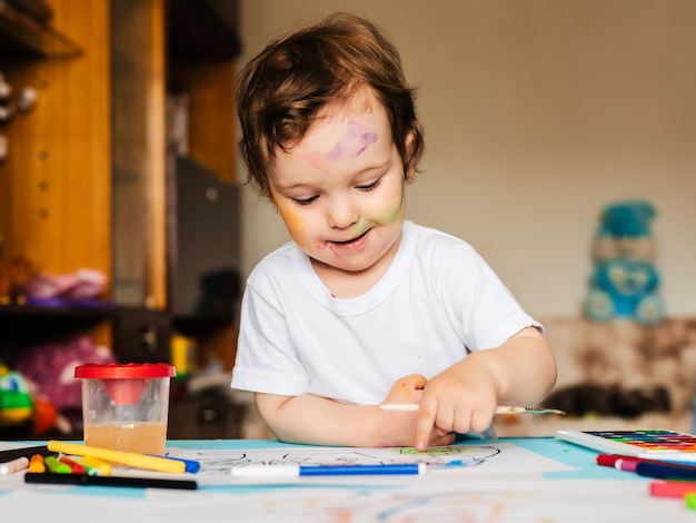 Um menino pequeno e bonito pinta com pincéis e tintas coloridas em uma folha de papel