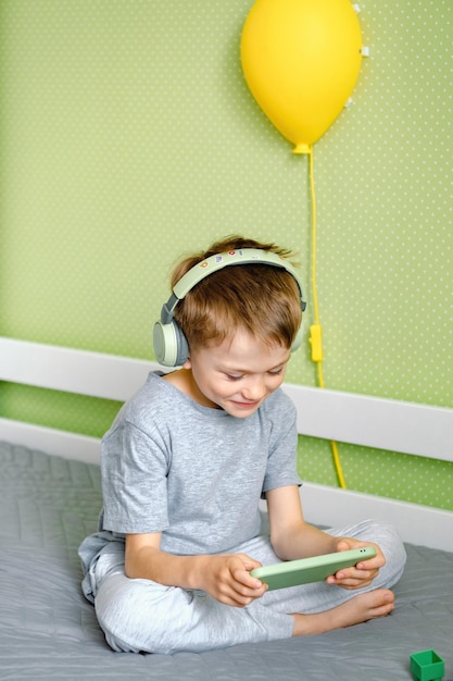 Um menino pré-escolar sentado na cama usando fones de ouvido sem fio e usando telefone celular