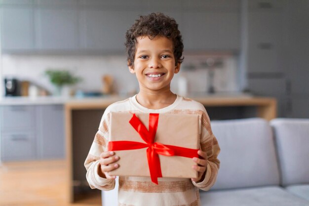 Um menino preto feliz segurando orgulhosamente um presente embrulhado com uma fita vermelha brilhante.