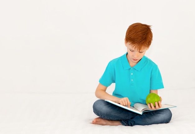 Um menino ruivo bonito está lendo um livro na cama e segurando uma maçã verde na mão