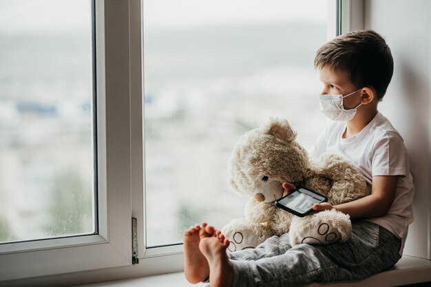 Um menino se senta em uma janela com um ursinho de pelúcia em quarentena e brinca em um telefone móvel. Prevenção de coronavírus e Covid - 19