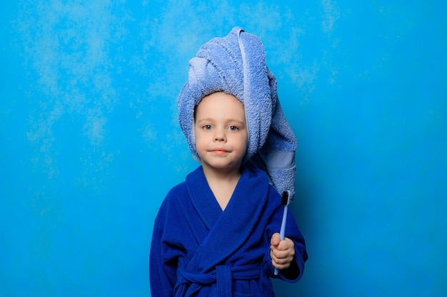 Um menino segura uma escova de dentes com um roupão e uma toalha em volta da cabeça sobre fundo azul