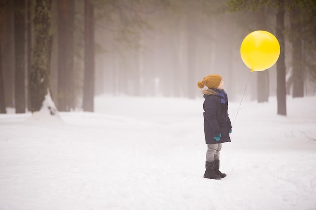 Um menino segurando um grande balão amarelo na floresta de inverno Aniversariante com balão