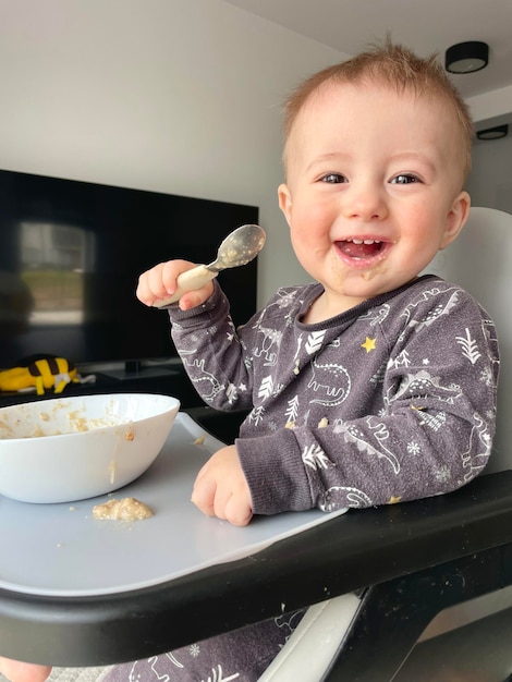 Foto um menino sentado em uma cadeira alta come mingau sozinho com uma colher