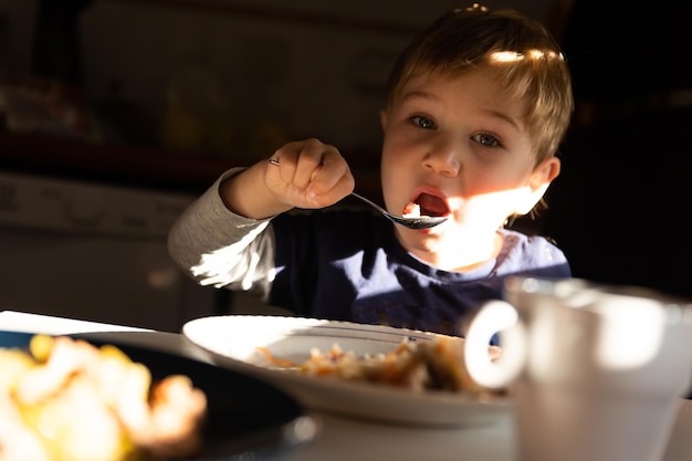 Um menino toma café da manhã na mesa come com uma colher e olha para a câmera