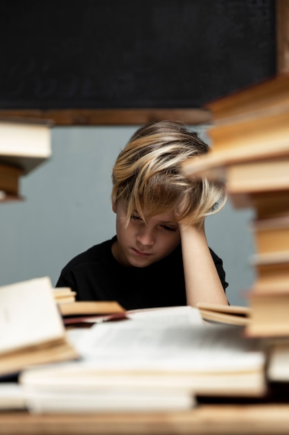 Um menino triste em uma camiseta preta está sentado a uma mesa com um monte de livros