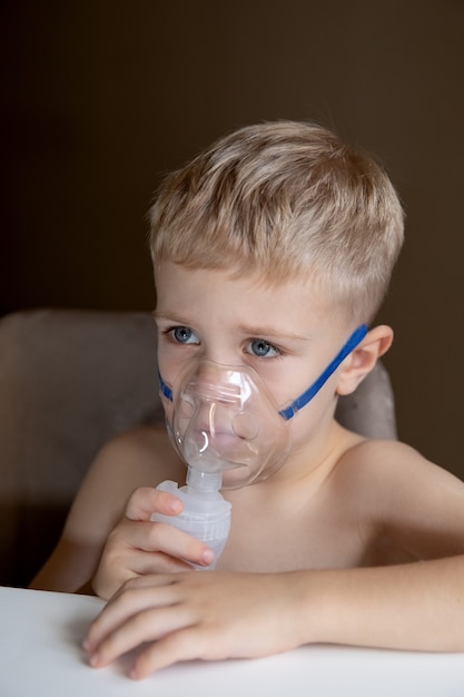Foto um menino triste faz inalações com um nebulizador em casa, ele está doente conceito de saúde