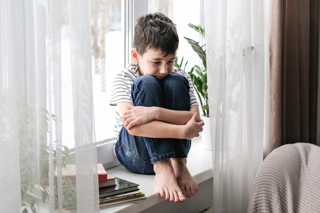 Um menino triste senta-se no parapeito da janela abraçando os joelhos depressão de mau humor o menino está triste sozinho em casa