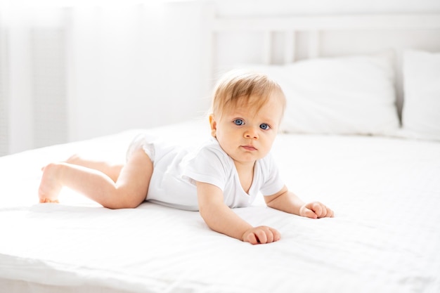 Foto um menino uma criança loira com olhos azuis em um bodysuit branco está em quatro patas em casa no quarto em uma cama com lençóis brancos e olha para a câmera