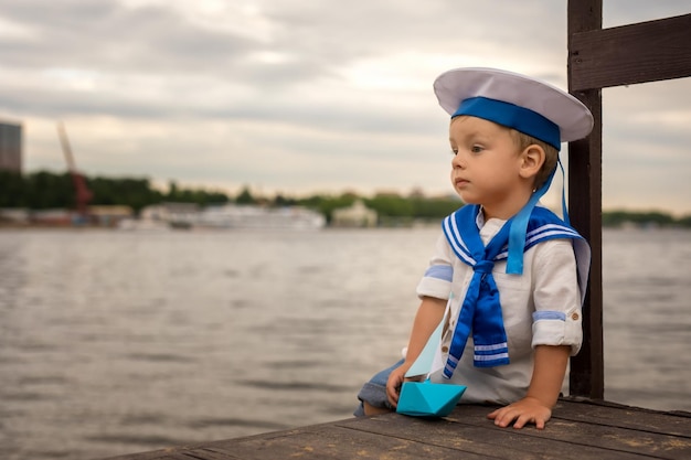Um menino vestido de marinheiro senta-se na praia com um barquinho de papel