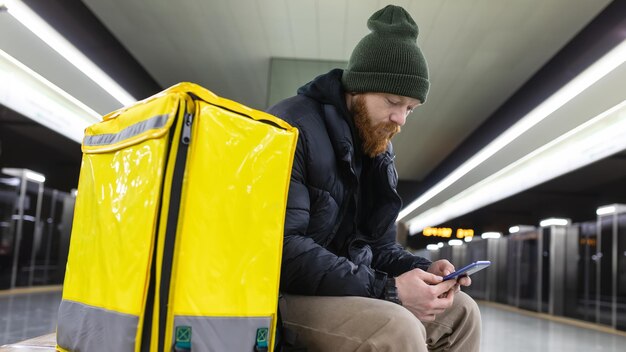 Um mensageiro com uma mochila amarela segura um celular e espera um trem do metrô na plataforma