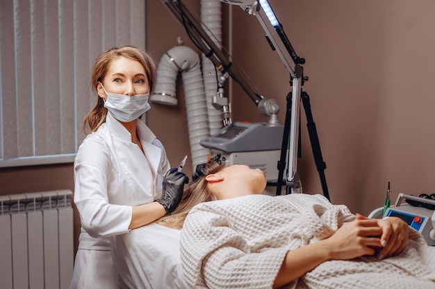 Um mestre cosmetologista em uma máscara branca protetora com uma seringa nas mãos e um cliente mentiroso no escritório de um salão de beleza