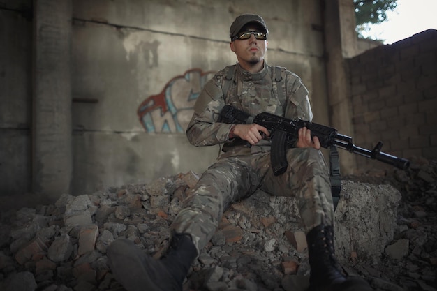 Um militar em um uniforme de camuflagem cinza usando um boné e óculos táticos com um grande átomo preto nas mãos está sentado no chão dentro de um grande prédio destruído