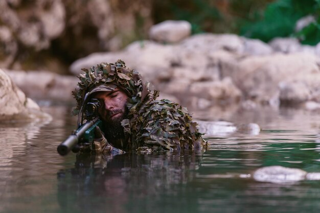 Foto um militar ou jogador de airsoft em um terno de camuflagem esgueirando-se no rio e apontando de um rifle de atirador para o lado ou para o alvo