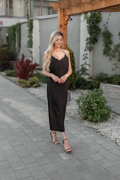 Um modelo de mulher elegante, feliz e elegante, com sorriso em um vestido preto longo e elegante caminha na rua