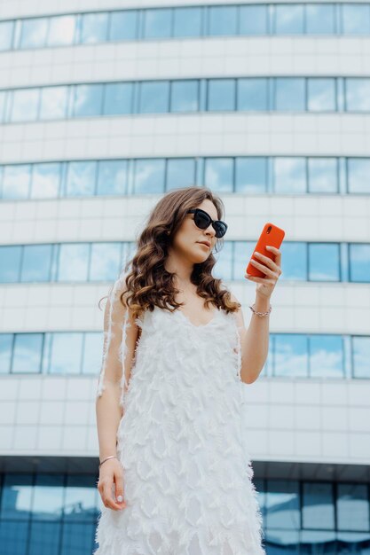 Um modelo elegante posa com um telefone do lado de fora Empresária de vestido branco e óculos de sol no fundo do escritório