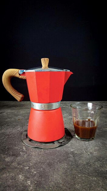 Foto um mokapot vermelho e um pequeno copo cheio de café estão prontos para serem servidos em uma mesa