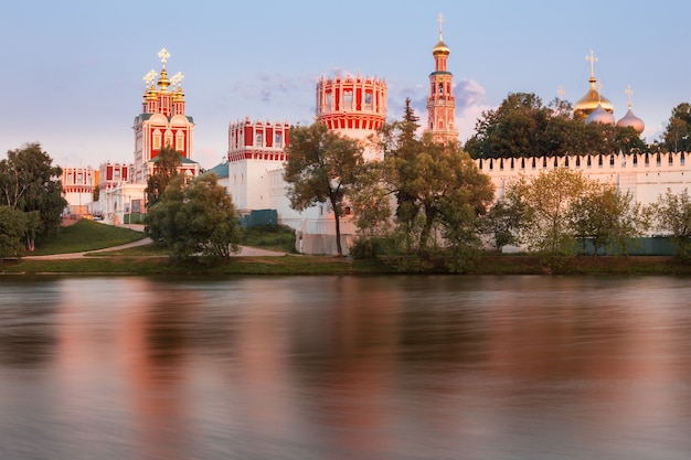 Um mosteiro ortodoxo russo com torres e igrejas de paredes caiadas de branco fica acima de um rio