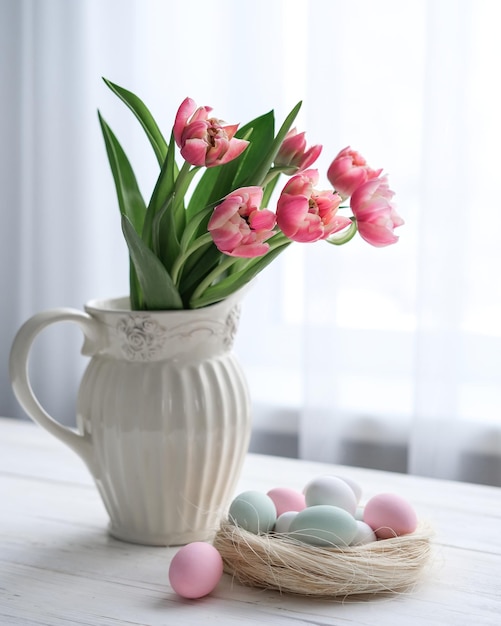 Um ninho com ovos em tons pastel suaves e um vaso com tulipas em uma mesa perto da janela