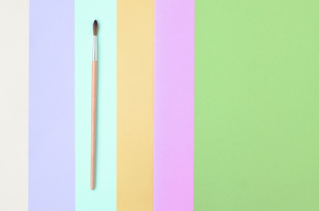 Um novo pincel fica sobre papel rosa, azul, verde, amarelo, violeta e bege