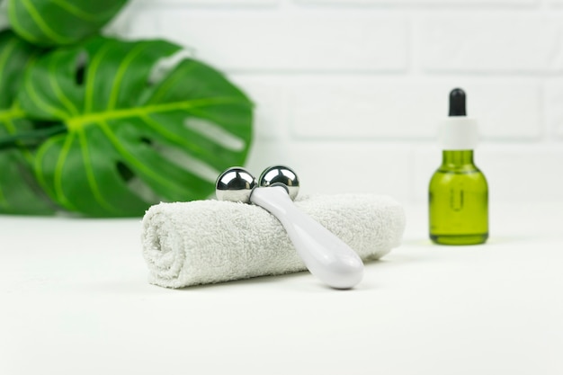 Um óleo verde CBD, um rolo para massagem facial, uma toalha de algodão branco e folhas verdes de monstera ficam em uma mesa branca em um banheiro