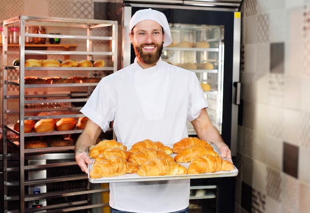 Um padeiro masculino considerável novo prende uma bandeja com croissants franceses na frente de uma padaria e sorri.