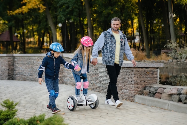 Um pai ajuda e ensina seus filhos pequenos a andar de Segway no parque durante o pôr do sol. Férias em família no parque.