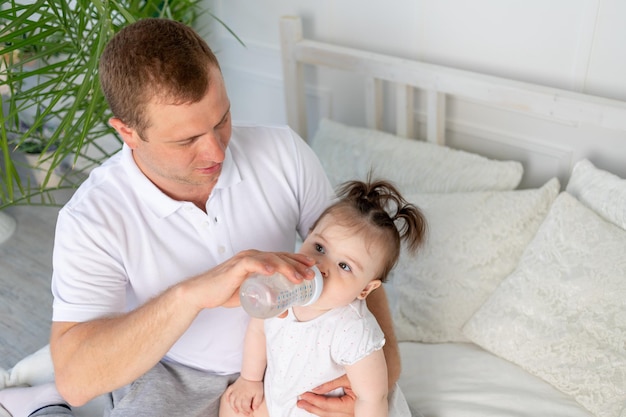 Um pai alimenta sua filhinha de uma garrafa em uma cama branca em casa o conceito de comida para bebê e cuidados infantis