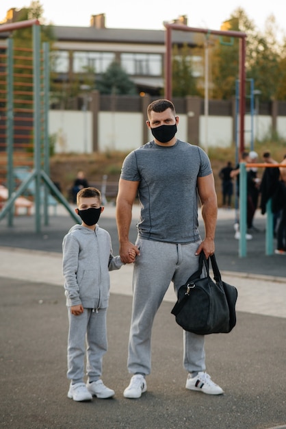 Um pai e um filho estão em um campo de esportes com máscaras após o treino durante o pôr do sol.