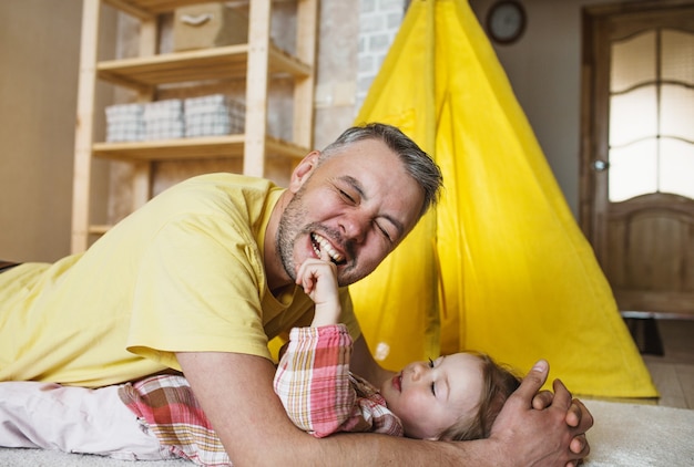 Um pai feliz morde o dedo de sua filha enquanto brincam juntos no chão em casa. jogos conjuntos do pai com o filho