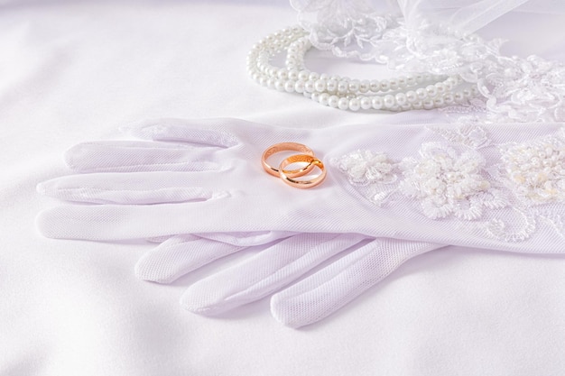 Um par de anéis de casamento de ouro deitam-se em lujosas luvas brancas de casamento fundo de cetim branco com pérolas conceito de acessórios de casamento
