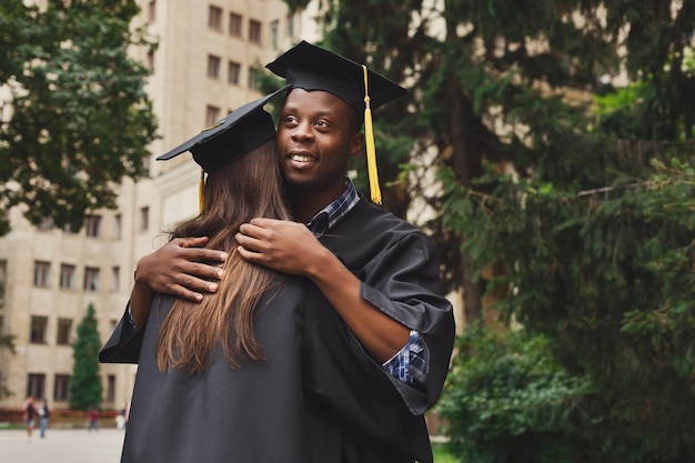 Um par de estudantes multiétnicas abraçados na formatura. Educação, qualificação e conceito de vestido.