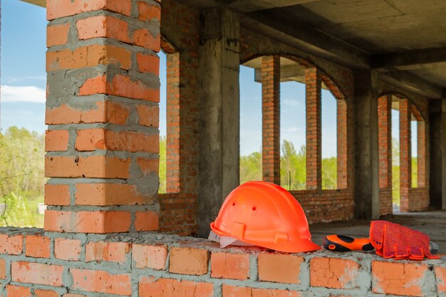 Foto um par de luvas e um capacete de pedreiro no canteiro de obras de uma casa de tijolos vermelhos.